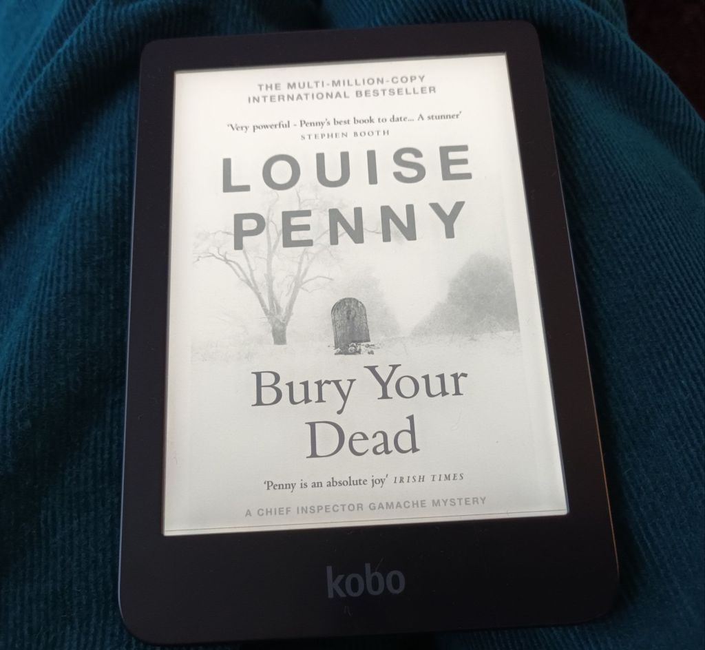 My e-reader showing the cover of 'Bury Your Dead' by Louise Penny. It features a snowy scene with a tombstone. 